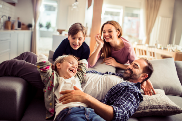 A smiling and laughing family of four plays together on a living room couch.
